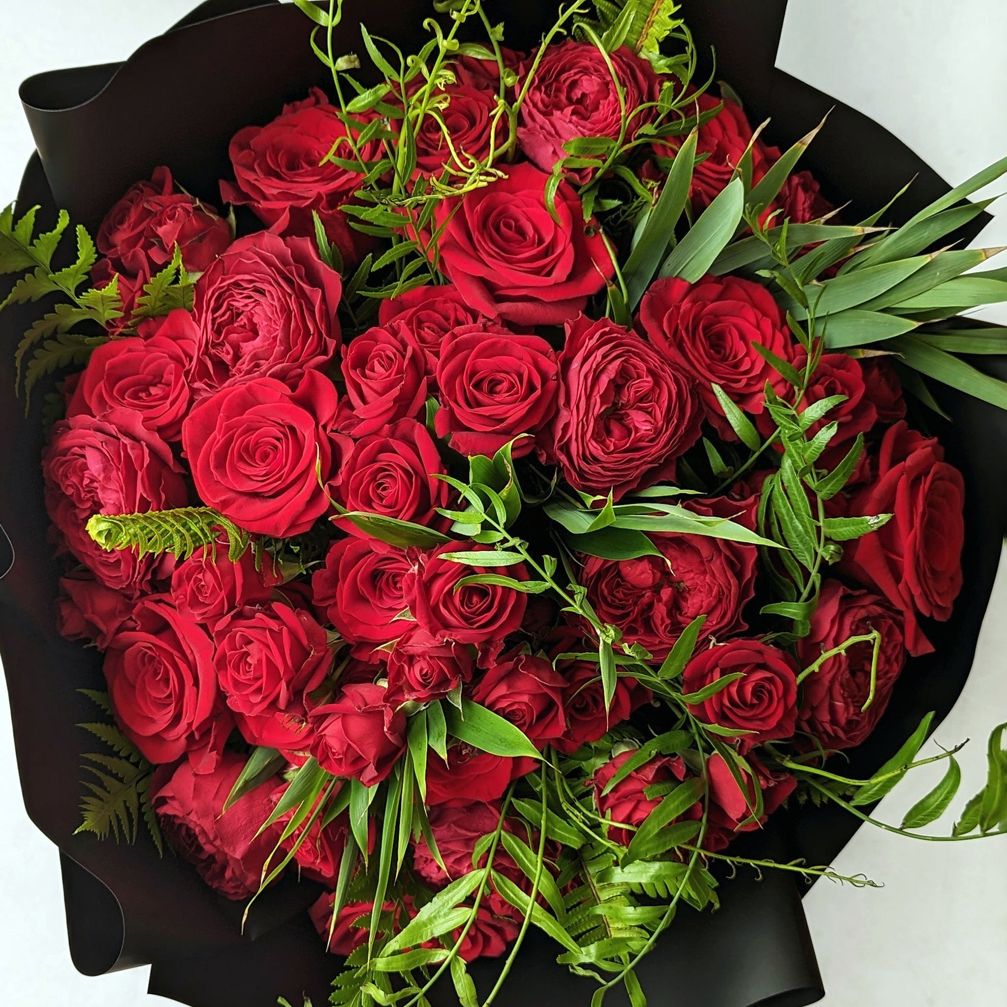 Carmine Bouquet, consisting of long single stem red spray roses of two varieties, with red spray roses, tied in with seasonal green foliage.